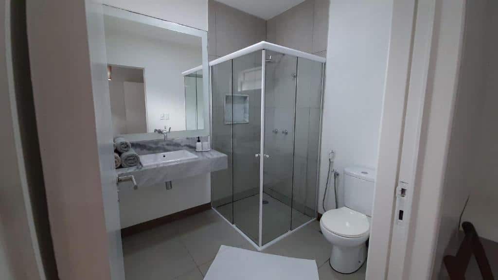 Banheiro da Pousada Recanto Ha Mar com um box de vidro, pia com toalhas, espelho e um vaso sanitário