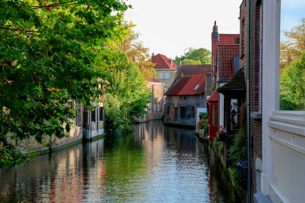 Cena serena de um canal em Bruges, conhecida por seus cursos d'água. Casas tradicionais com fachadas de tijolos e telhados vermelhos distintos margeiam o canal, refletindo sua charmosa arquitetura na água calma, mas há folhagem verde no entorno, adicionando um toque de natureza à paisagem urbana.
