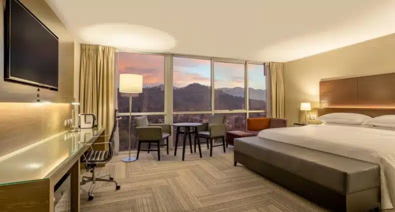 Quarto do Sheraton Santiago Hotel & Convention Center, com cama de casal do lado direito da imagem no centro do quarto do lado esquerdo uma mesa com duas cadeiras ao lado de uma janela panoramica com vista para a cidade e em frente a cama uma cômoda com TV. Representa hotéis perto do Cerro San Cristóbal em Santiago.