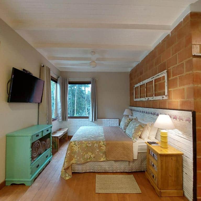 Quarto da Casa no Céu com uma cama de casal, mesa de cabeceira de madeira com abajur, uma TV montada na parede, um banco de madeira perto da janela e uma cômoda verde.