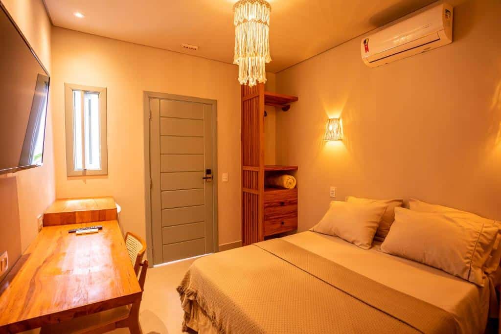 Quarto da Casa Savana. Uma cama de casal com um ar-condicionado e um abajur em cima. De frente uma mesa de trabalho, uma televisão e um frigobar. No fundo a pequena janela, a porta e um mini guarda-roupa. Foto para ilustrar post sobre pousadas pet friendly em São Paulo.