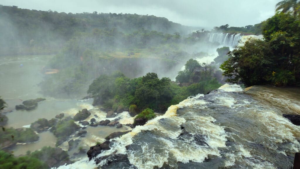Vista das Cataratas do Iguaçu em um dia nublado, com várias quedas d'água visíveis ao longe e uma névoa densa que cobre a paisagem. O rio corre com força entre as pedras, cercado por uma vegetação densa e verdejante.