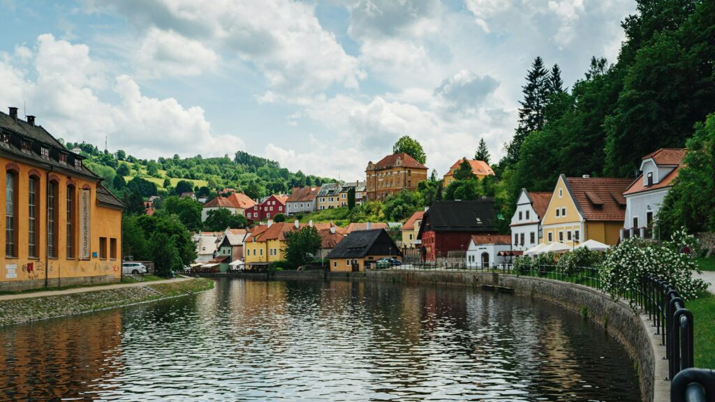 A pitoresca vila europeia de Český Krumlov, cortada por um rio. As casas coloridas com telhados de diferentes tons alinham-se ao longo da margem, destacando-se contra um cenário de colinas verdes arborizadas. O céu parcialmente nublado acima adiciona um toque dinâmico à cena tranquila. Uma calçada com uma grade preta curva-se ao longo do rio, oferecendo um local ideal para passeios relaxantes.