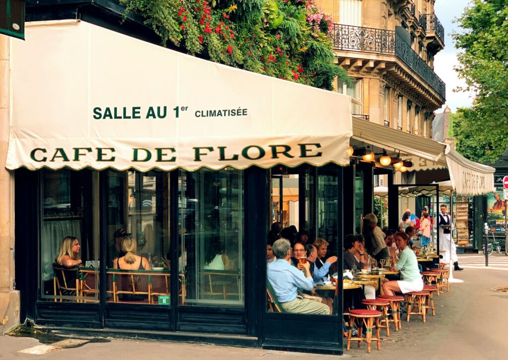 Fachada do Café de Flore, uma cafeteria com mesas na calçada em Saint Germain. Várias pessoas estão sentadas ali degustando de bebidas e comidas durante o dia. - Foto: Chahriar Hariri via Unsplash
