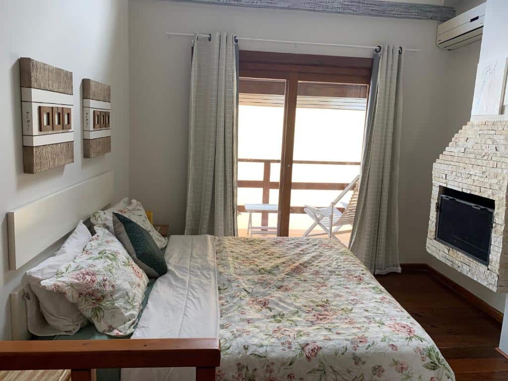 Foto de um quarto do chalé aconchegante para a família no Centro de Penedo. Tem uma cama box de casal no centro, e nós vemos ela de lado. Na frente dela há uma lareira. Ao lado da cama, no centro da parede, há uma porta de vidro que dá acesso à uma varanda.