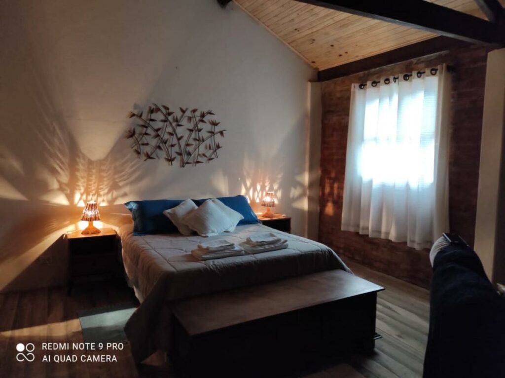 Quarto do Chalé Lavanda com uma cama de casal no meio e entre duas mesinhas com abajures. Em frente a cama está um banco e um sofá. Ao seu lado direito está uma janela com cortina. A foto faz parte da lista de melhores chalés em São Francisco Xavier.