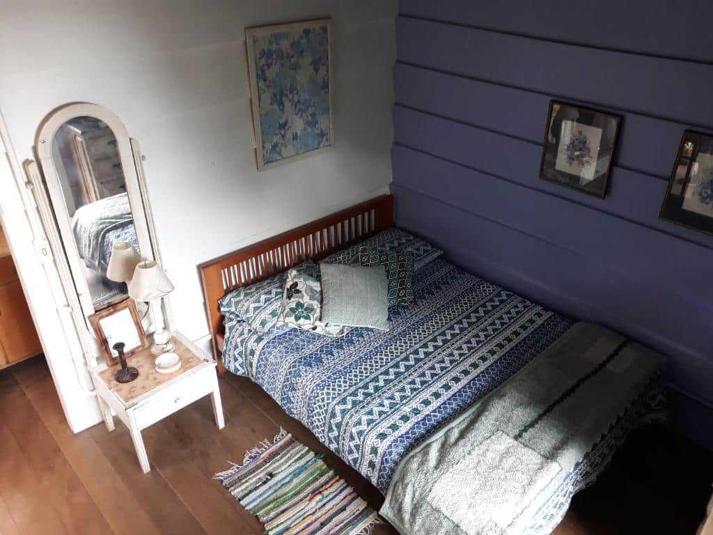 Quarto do Chales Bamboo Jungle com uma cama de casal, roupa de cama estampada, dois travesseiros, uma cabeceira de madeira, um criado-mudo com um abajur, um espelho de parede e uma obra de arte emoldurada.