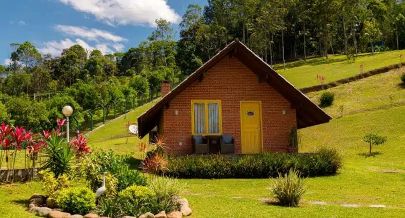 Fachada de um dos Chalés Pomerode que mostra um chalé de parede de tijolos, com uma porta e uma janela, e duas cadeiras. Em volta do chalé tem um amplo gramado verde com plantas e árvores em volta. Imagem ilustrando post Chalés em Pomerode.