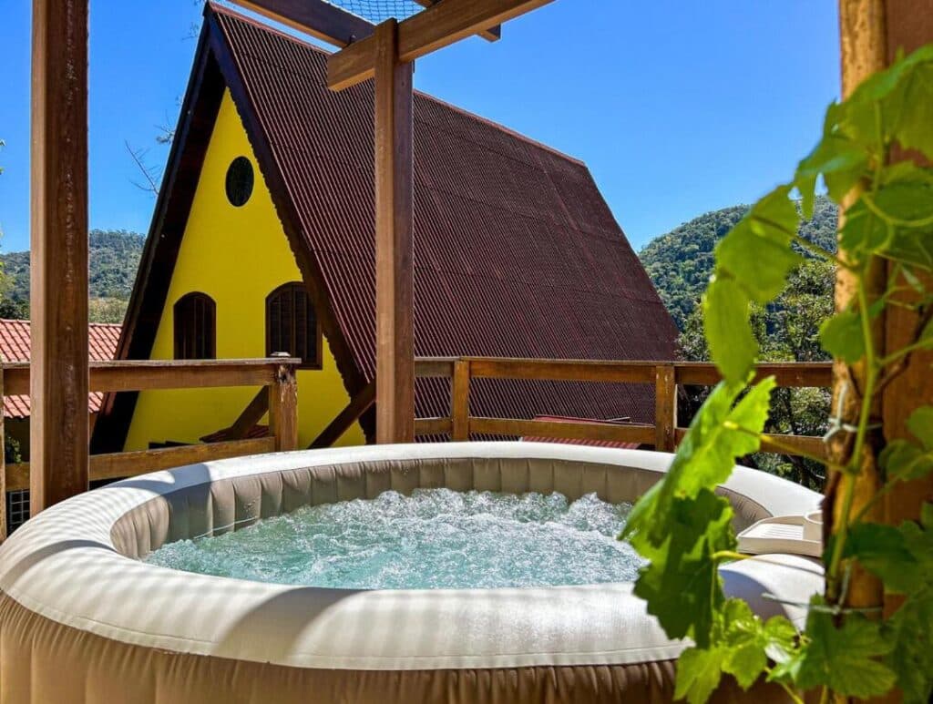 Banheira de hidromassagem em um deck de madeira com vista para uma casa amarela em formato de A e colinas verdejantes ao fundo. A foto pertence ao Charmoso chale, em Itaipava.