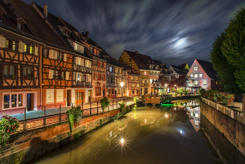 Esta imagem noturna captura a atmosfera encantadora da cidade de Colmar, na França. Mostra um canal ladeado por casas de enxaimel coloridas e ricamente iluminadas, refletindo suas luzes na água tranquila. As nuvens rápidas no céu e a lua visível criam um fundo dramático. A cena é acentuada por flores vivas nas margens e luzes cintilantes que destacam a arquitetura tradicional e o ambiente romântico da cidade.