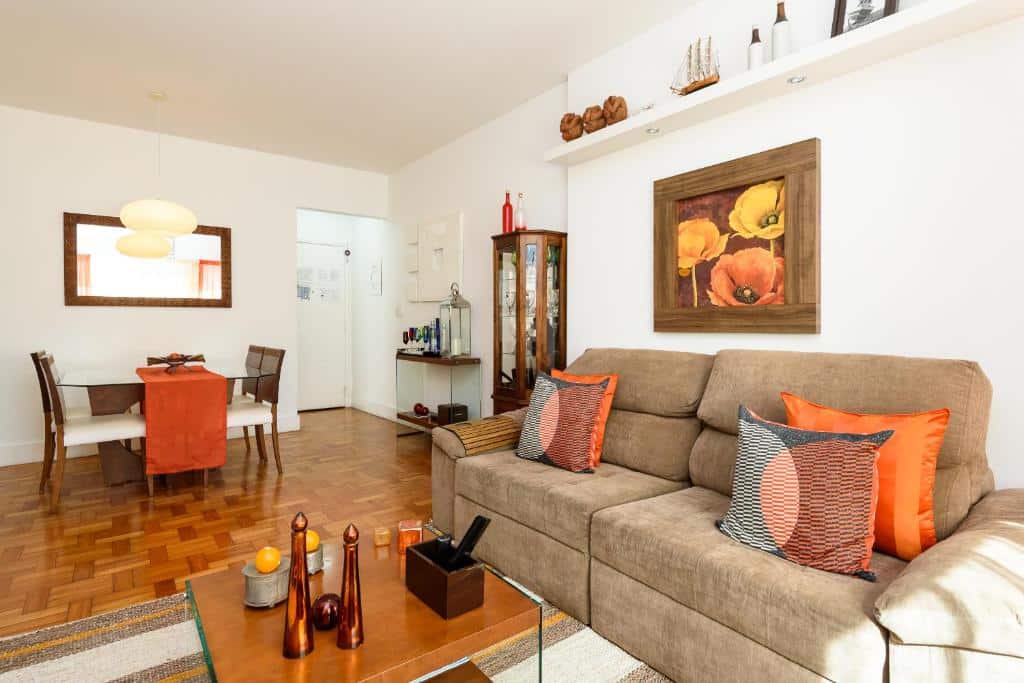 Uma sala no Copacabana Beach Amazing Apartment. Mostra um sofá na direita, uma mesa de centro à sua frente. No fundo à esquerda há uma mesa com cadeiras para o jantar. O espaço é todo decorado em tons marrons e laranja. As paredes são brancas e o piso é de madeira. 