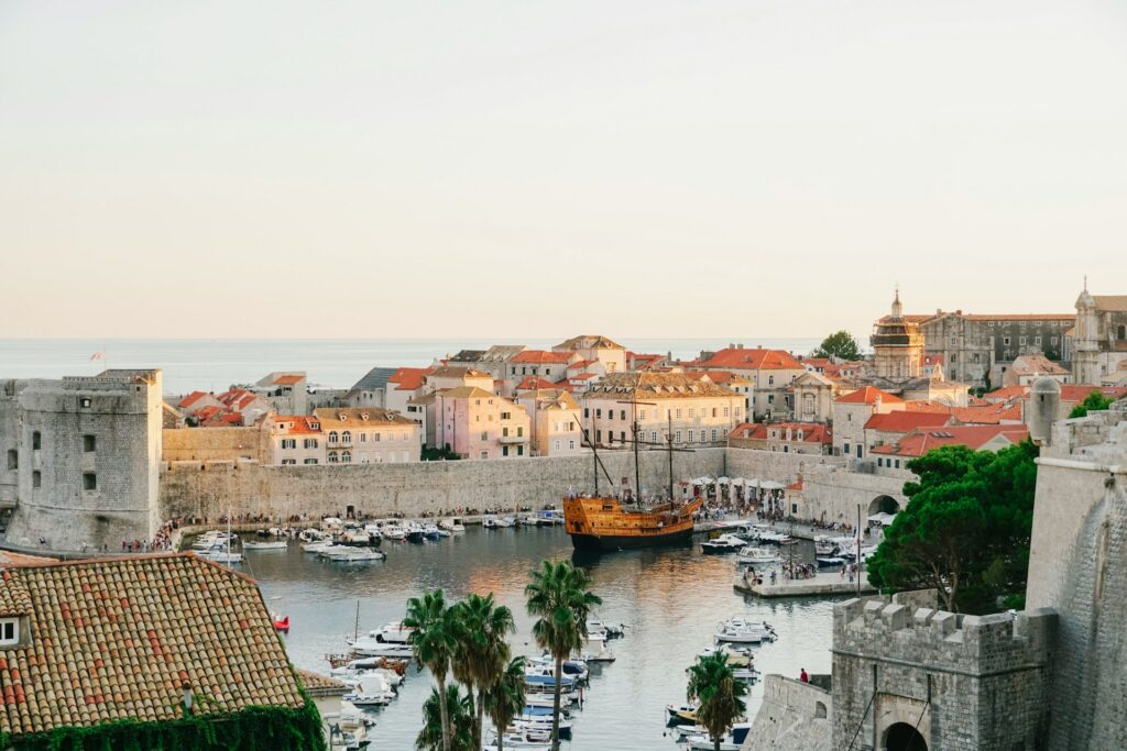 Antigo porto da cidade de Dubrovnik, Croácia, ao entardecer, com barcos ancorados, incluindo uma réplica de um antigo navio à vela. A cidade é cercada por suas famosas muralhas de pedra, e os edifícios com telha vermelha destacam-se contra o fundo do mar Adriático. A cena é complementada por uma suave iluminação dourada do sol poente, realçando a beleza da atmosfera histórica do local.