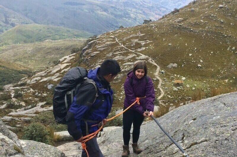 Duas pessoas escalando uma pedra gigante, com vista atrás de natureza e montanhas e uma trilha longa no meio delas. Representa o que fazer no Peru