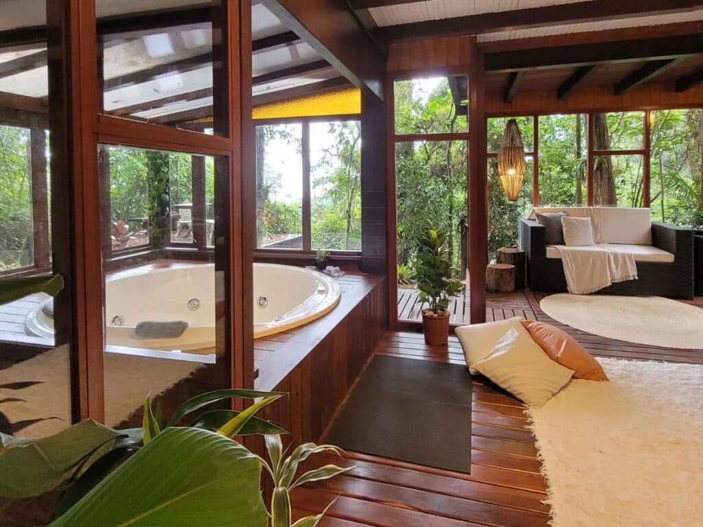 Sala do Espaço Verde Novo Chalés com banheira de hidromassagem no lado esquerdo, área de estar, janelas grandes de vidro, plantas verdes e iluminação suave.