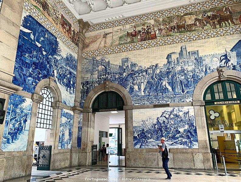 Imagem de dentro da Estação Ferroviária de São Bento com uma pessoa no centro com câmera fotográfica o lugar tem parede revestidas com azulejos decorados com a história do país. Representa o que fazer no Porto.