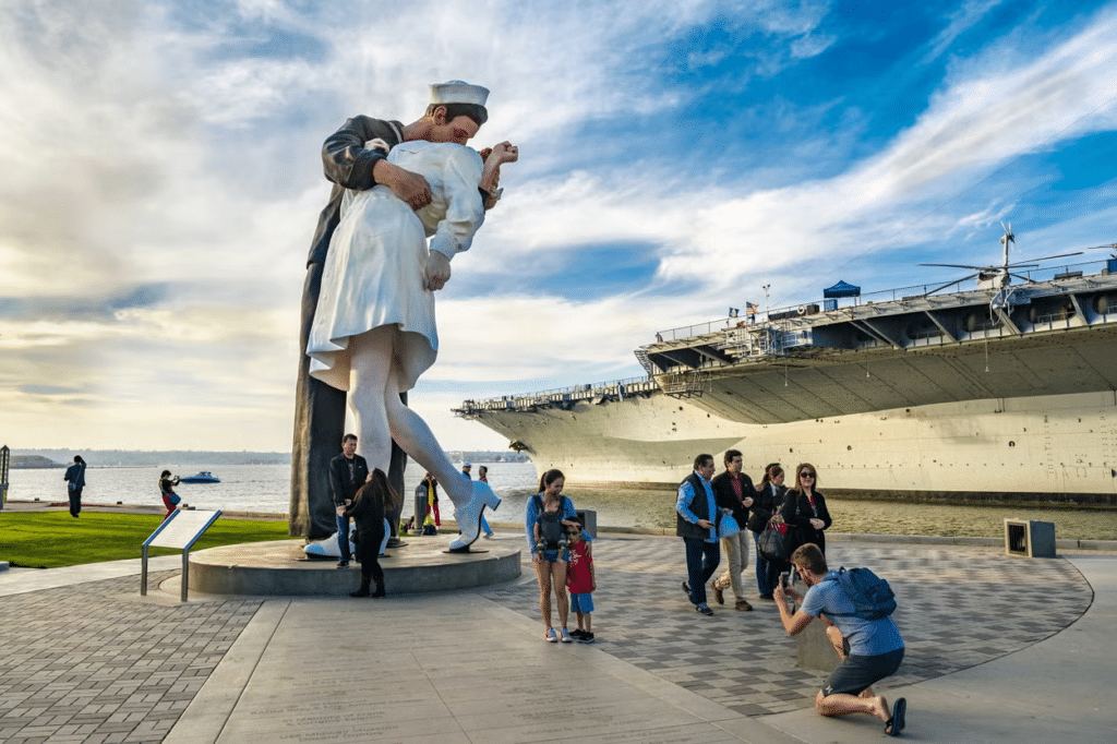 Uma estátua de um casal se beijando, pessoas ao redor tirando foto e no fundo um navio, no museu USS Midway.