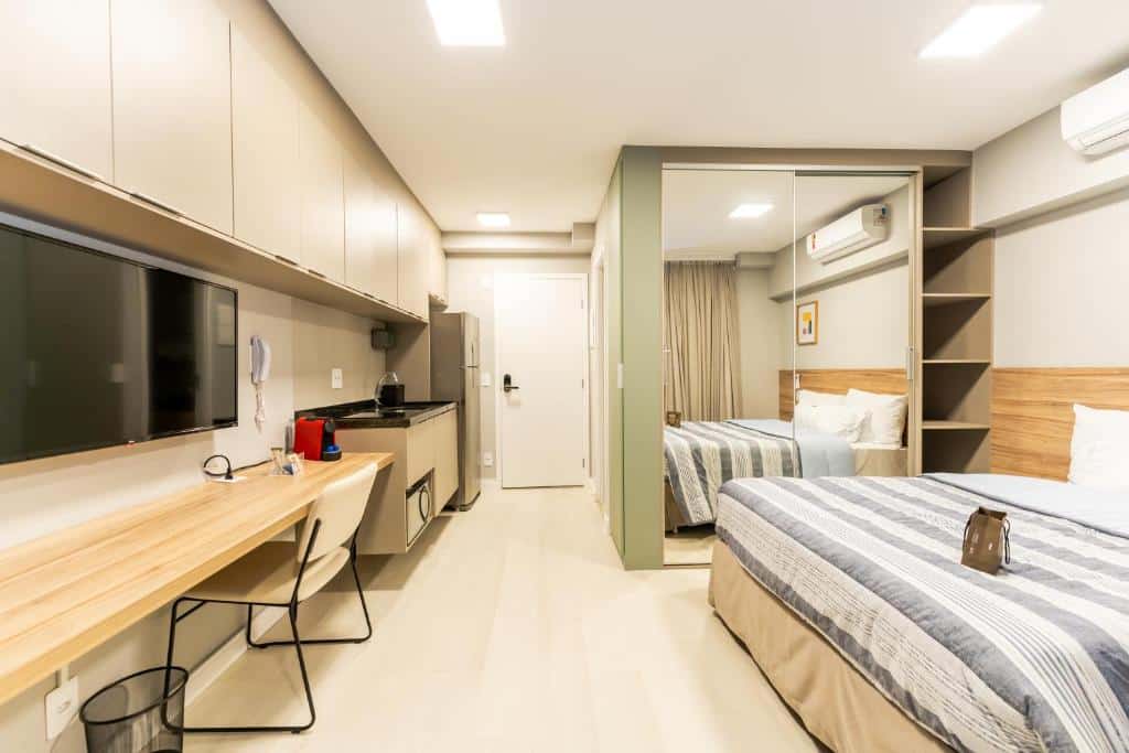 A imagem retrata o interior de um dos estúdios do Apartamentos Stdudio Allure Moena, com uma cozinha compacta à esquerda. A cozinha tem armários acima e abaixo da bancada, onde há uma cafeteira e itens de cozinha. Uma TV plana está fixada na parede acima da bancada que tem uma cadeira à frente. À direita, há uma cama de casal com roupa de cama listrada em cinza, com cabeceira de madeira. Há um espelho de corpo inteiro ao lado da cama mais distante. No fundo, vê-se uma porta fechada, indicando a entrada no estúdio. O piso é claro e as paredes são brancas, criando uma atmosfera clara e acolhedora.