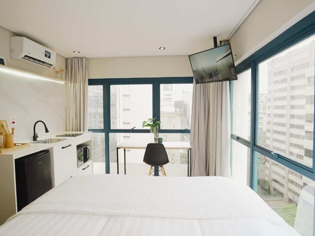 Foto do interior do estúdio do Apê Paulista Bela Cintra, um dos airbnb em São Paulo, sendo ele bem iluminado com uma grande janela que revela uma vista da cidade. A cama de casal está centralizada no espaço, com roupa de cama branca. À esquerda, há uma cozinha compacta com armários brancos, uma pia e um micro-ondas. À direita, uma mesa pequena com cadeira e uma TV de tela plana fixada na parede acima. A decoração inclui uma grande arte abstrata na parede e um ar-condicionado no alto, acima da cama.