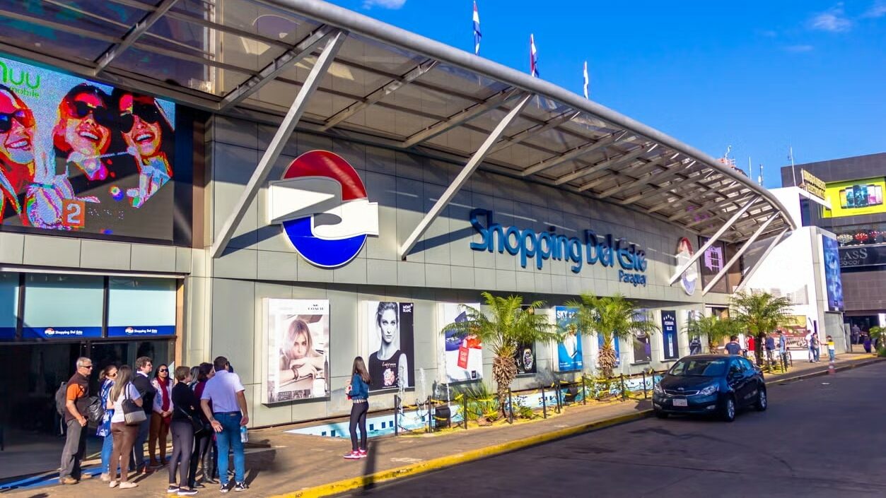 Fachada do Shopping Del Este, localizado no Paraguai. A entrada do shopping é moderna, com um grande logotipo circular vermelho e azul. Várias pessoas estão reunidas na frente do shopping, e ao fundo, há anúncios e vitrines de lojas.