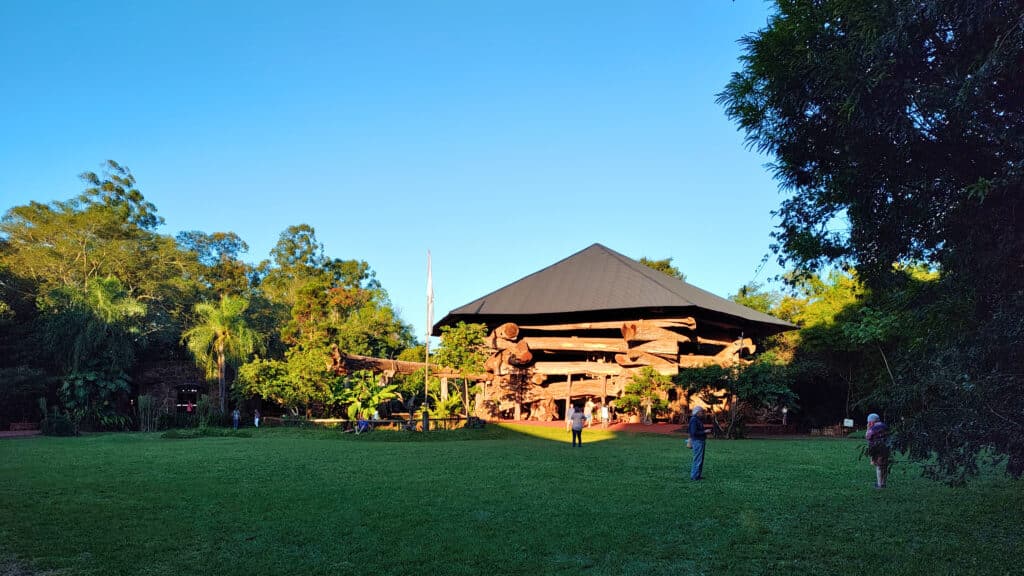 A estrutura principal, ao centro da imagem, é feita de troncos de madeira robustos e possui um telhado triangular inclinado. O local é cercado por um gramado amplo e vegetação densa, com algumas pessoas caminhando pela área e um céu azul. sem nuvens em Puerto Iguazu.