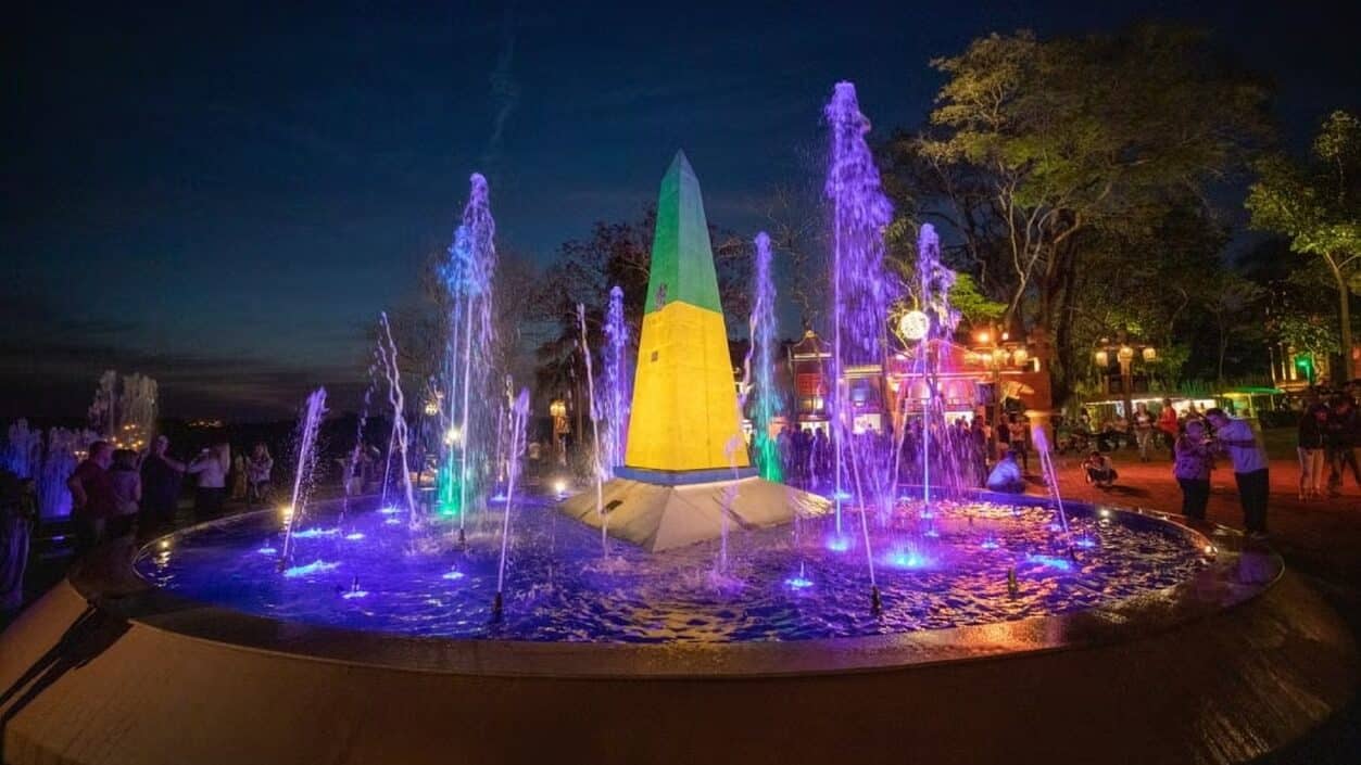 Fonte luminosa no Marco das Três Fronteiras em Foz do Iguaçu, iluminada à noite com luzes coloridas. A estrutura central, em forma de obelisco com as cores do Brasil, é cercada por jatos d'água que mudam de cor. Ao fundo, pessoas passeiam e apreciam a atração.
