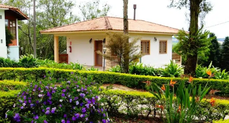 A imagem mostra uma charmosa construção do Hotel Fazenda Itáytyba no Paraná, cercada por belos jardins bem cuidados. A casa é de cor clara, com telhado de telhas vermelhas e janelas de madeira. O jardim na frente da casa é adornado com diversas plantas e flores coloridas, como arbustos floridos de tom roxo e flores exóticas laranja, além de cercas vivas bem podadas que delineiam os canteiros. Ao fundo, árvores altas completam a paisagem natural, dando ao ambiente uma sensação de tranquilidade e proximidade com a natureza. O cenário transmite uma atmosfera serena e acolhedora, ideal para relaxar em meio ao verde.