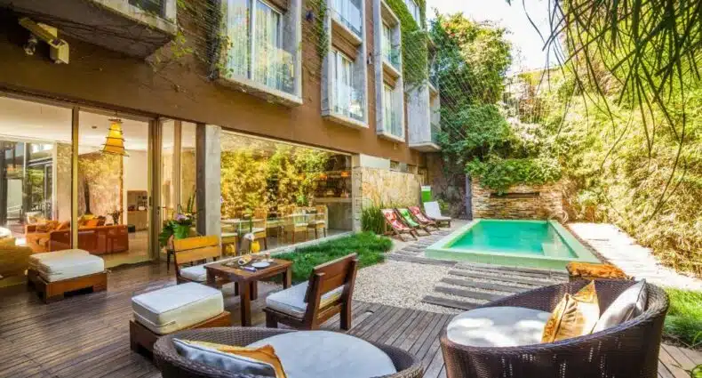 Pátio moderno do Mine Hotel, um dos hotéis românticos em Buenos Aires, com cadeiras de vime, deck de madeira, mesa de jantar, espreguiçadeiras e uma pequena piscina cercada por plantas.