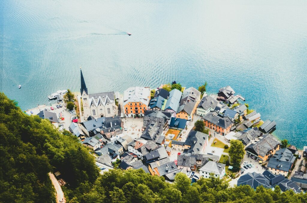 Vista aérea da cidade de Hallstatt, na Áustria, ao lado de um lago cristalino. A cidade é composta por casas coloridas com telhados de ardósia, intercaladas por ruas estreitas e uma igreja proeminente com uma torre pontiaguda. Na água do lago pequenas embarcações são visíveis e árvores verdes cercam uma parte da cidade.