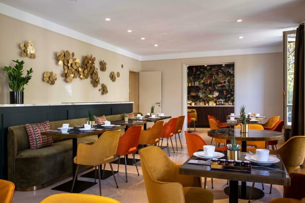 Salão de café da manhã do Hôtel Aiglon, onde várias mesas com cadeiras e sofás estão dispostas. Há utensílios e plantinhas nas mesas, e as paredes contam com decorações. Ao fundo, uma porta leva ao balcão com itens do café.