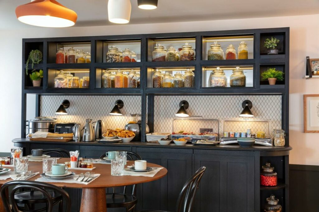 Bancada de café da manhã no Hotel Bastille Speria, onde ficam cereais, pães, frutas, iogurtes e outros itens. Logo em frente há uma mesa grande com cadeiras e utensílios.