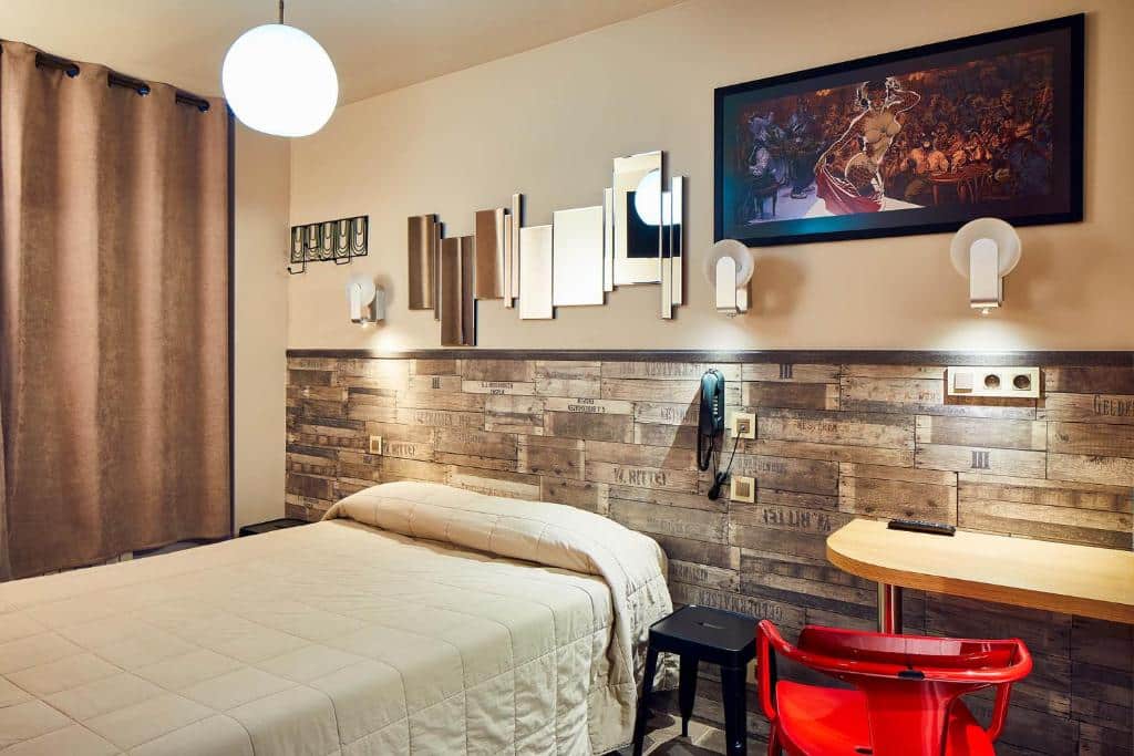 Quarto do Hôtel de Roubaix, um dos hotéis no Le Marais em Paris. Uma cama de casal está no centro do quarto e tem mesinhas de cabeceira e abajures dos dois lados. Ao lado esquerdo há uma janela com cortinas, enquanto do lado direito fica uma escrivaninha com cadeira e luminária. Há decorações na parede, como espelhos e quadros.