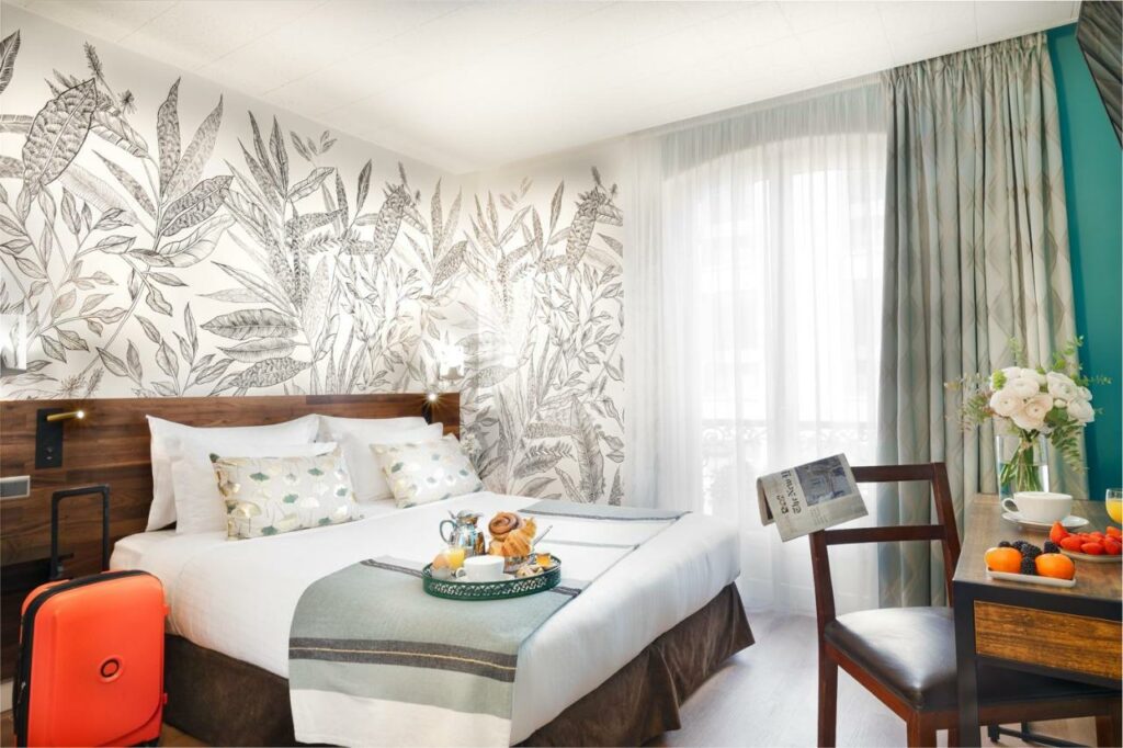 Quarto do Hôtel Delavigne, um dos hotéis 3 estrelas em Paris. Uma cama de casal está encostada na parede esquerda do local com mesinhas de cabeceira e abajures dos dois lados, assim como uma bandeja com itens de café da manhã aos pés. Logo em frente fica uma mesa com cadeira, jornal, vaso de flores e outros alimentos do café. Na lateral direita da cama há uma janela com cortinas.