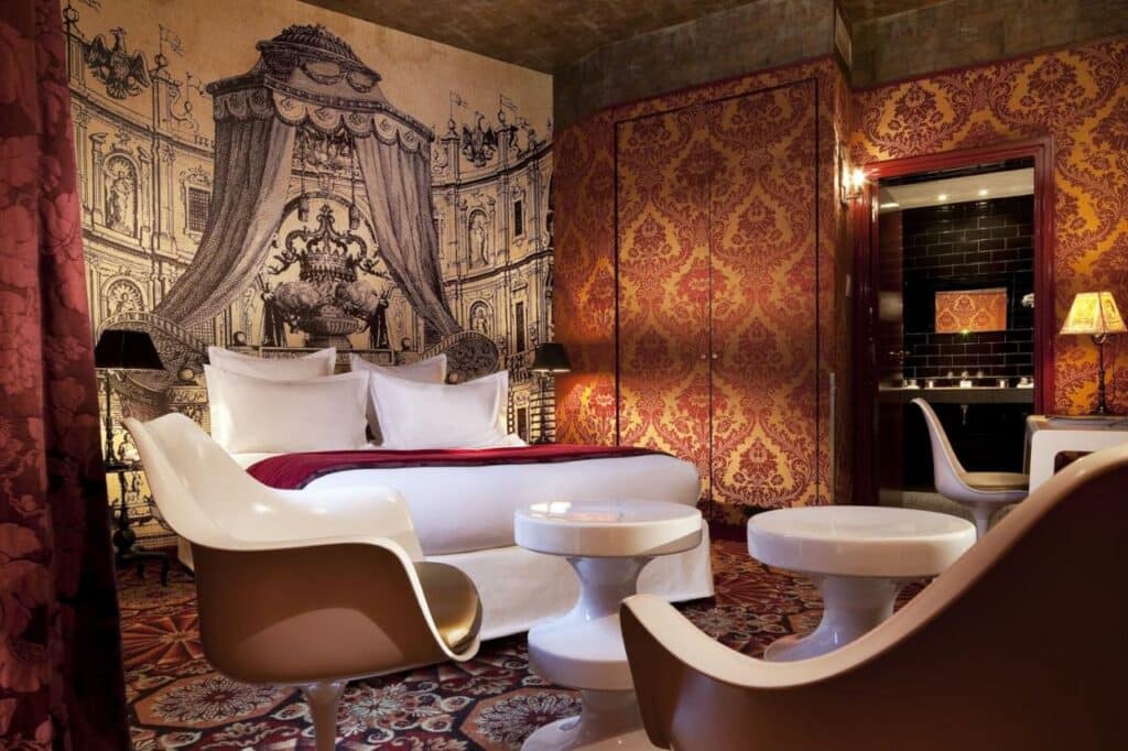 Quarto do Hotel du Petit Moulin, um dos hotéis 4 estrelas em Paris. Uma cama de casal redonda está ao lado esquerdo do quarto e tem mesinhas de cabeceira e abajures dos dois lados, assim como um guarda-roupas ao lado esquerdo. Logo em frente há uma área de estar com cadeiras e mesinhas, e ao lado fica uma escrivaninha com cadeira e abajur.