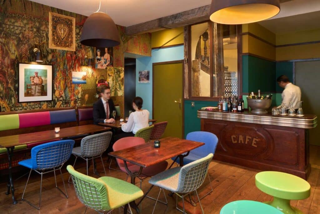 Pessoas sentadas na sala de refeições do Hotel du Petit Moulin, onde várias mesas com cadeiras e sofás coloridos estão dispostos. Há uma bancada na lateral direita onde um homem prepara bebidas.
