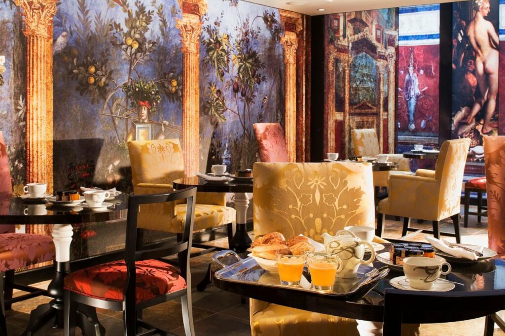 Salão de café da manhã do Hôtel Le Bellechasse Saint-Germain, onde várias mesas e cadeiras estão dispostas ao redor. Há quadros e papéis de parede no local.