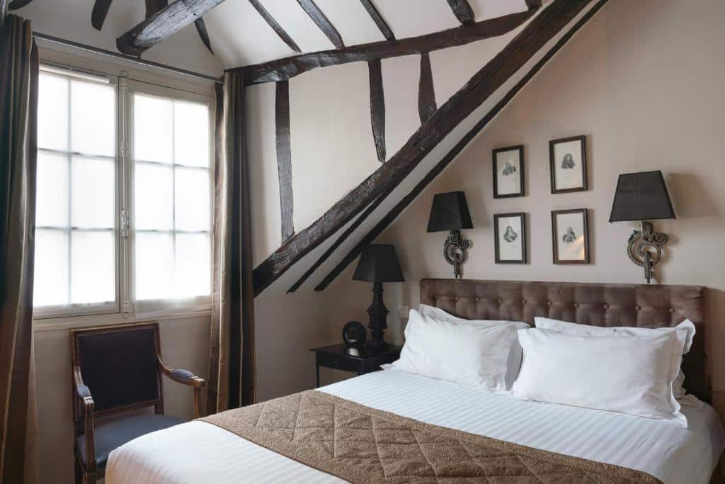 Quarto do Hotel Saint-Louis Marais, um dos hotéis 3 estrelas em Paris. Uma cama de casal está ao lado direito do quarto e tem mesinhas de cabeceira e abajures dos dois lados, assim como quadrinhos na parede acima. Ao lado esquerdo há uma poltrona em frente a uma janela com cortinas.