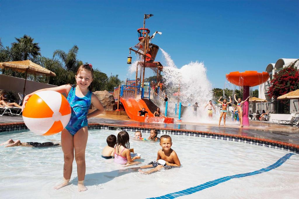 Área de lazer do Howard Johnson by Wyndham Anaheim. Crianças brincando na piscina rasa, atrás um brinquedo aquático. Imagem para ilustrar post sobre hotéis perto da Disney Califórnia.