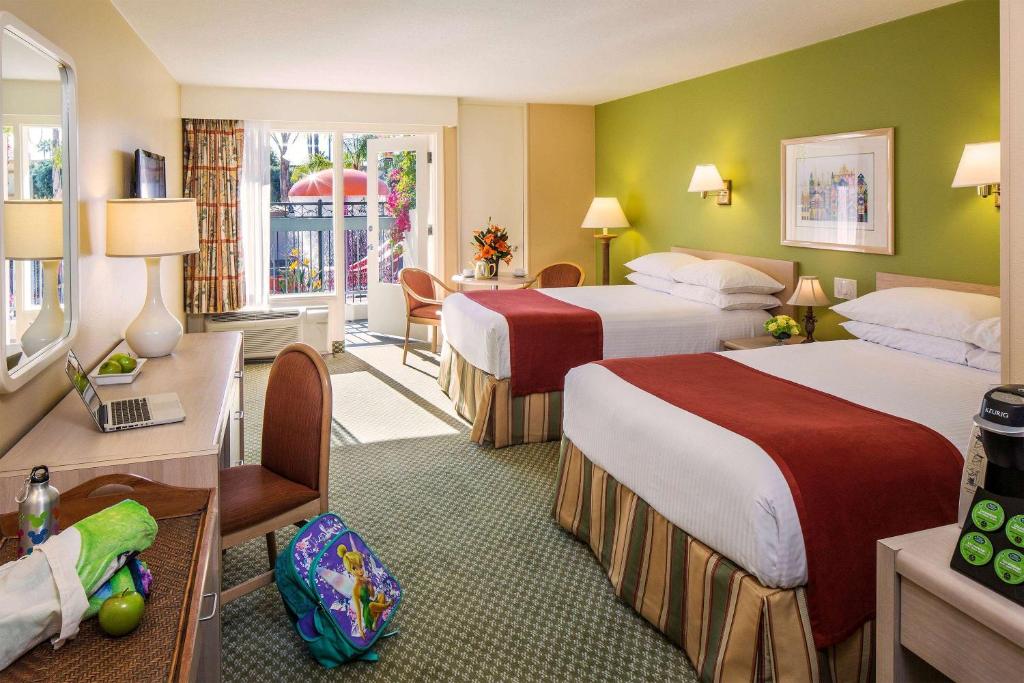 Quarto do Howard Johnson by Wyndham Anaheim. Duas camas de casal do lado direito, entre elas e no fundo abajur. No fundo uma mesa com duas cadeiras e a porta da varanda aberta. Do lado esquerdo uma mesa de trabalho, um espelho, um abajur, uma mochila infantil e uma TV.