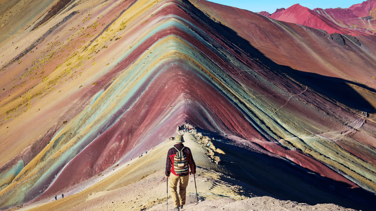 Homem de mochila e segurando dois apoios para caminhada, andando em direção à Montanha Arco-íres, composta por várias cores. Representa o que fazer no Peru