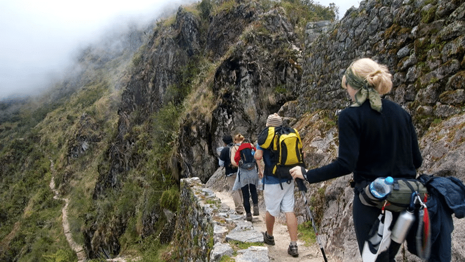 Pessoas caminhando em uma trilha com monstanhas em volta e muia natureza. Eles estão com mochilas e itens para fazer caminhada. Representa o que fazer no Peru