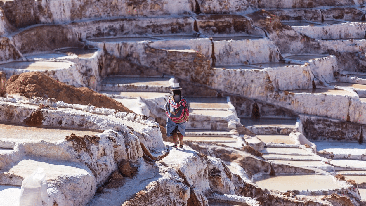 Uma mulher com roupas típicas do Peru em meio as salinas de Maras, durante o dia.