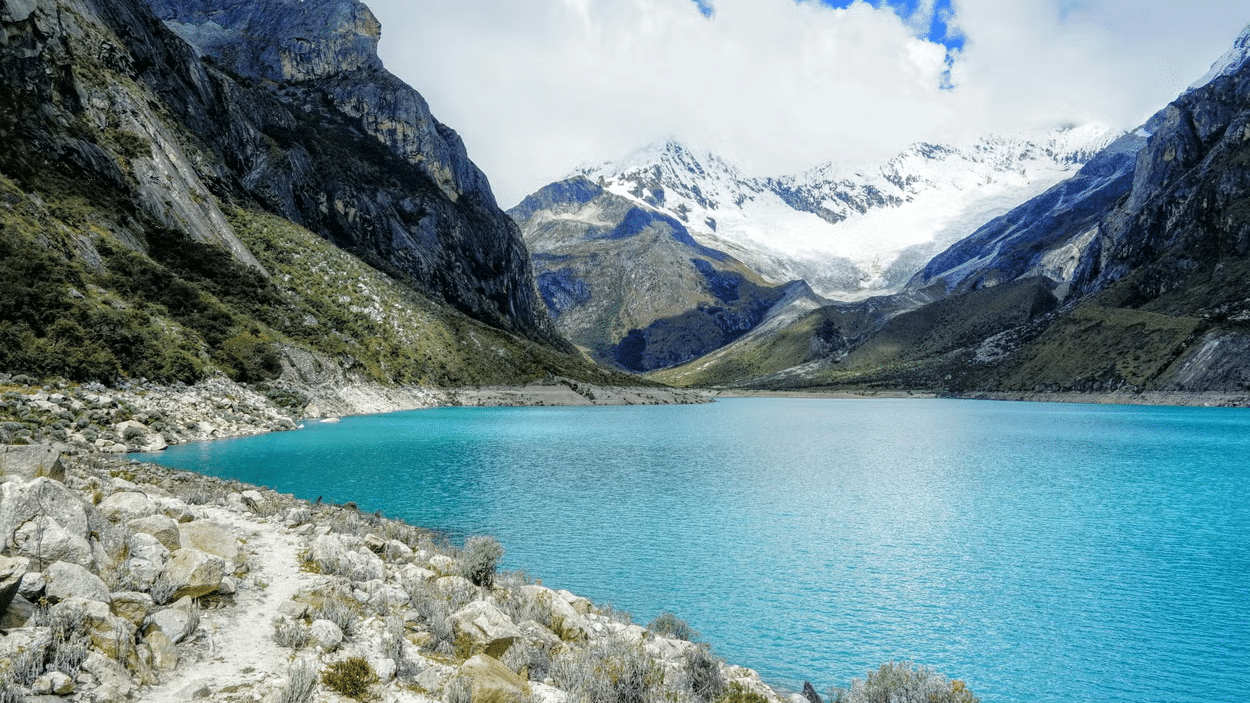 Lago azul cristalino com montanhas em volta cobertas de natureza, neve e núvens