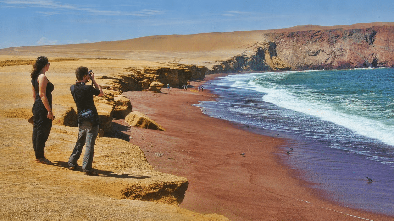 Duas pessoas viradas, de perfil, olhando uma praia que é rodeada por um deserto. Uma das pessoas está tirando foto, com a câmera no olho, segurando-a com as duas mãos. Foto: Civitiatis