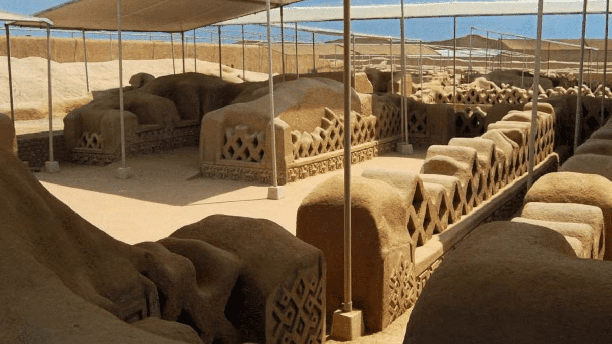 Esculturas feitas de barro, cobertas por uma estrututa de lona marrom, localizados em uma área extensa de terra, como se fosse um deserto. Representa o que fazer no Peru