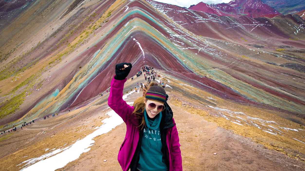 Mulher com um dos braços levantados, sorrindo, com montanhas coloridas atrás e pessoas andando.