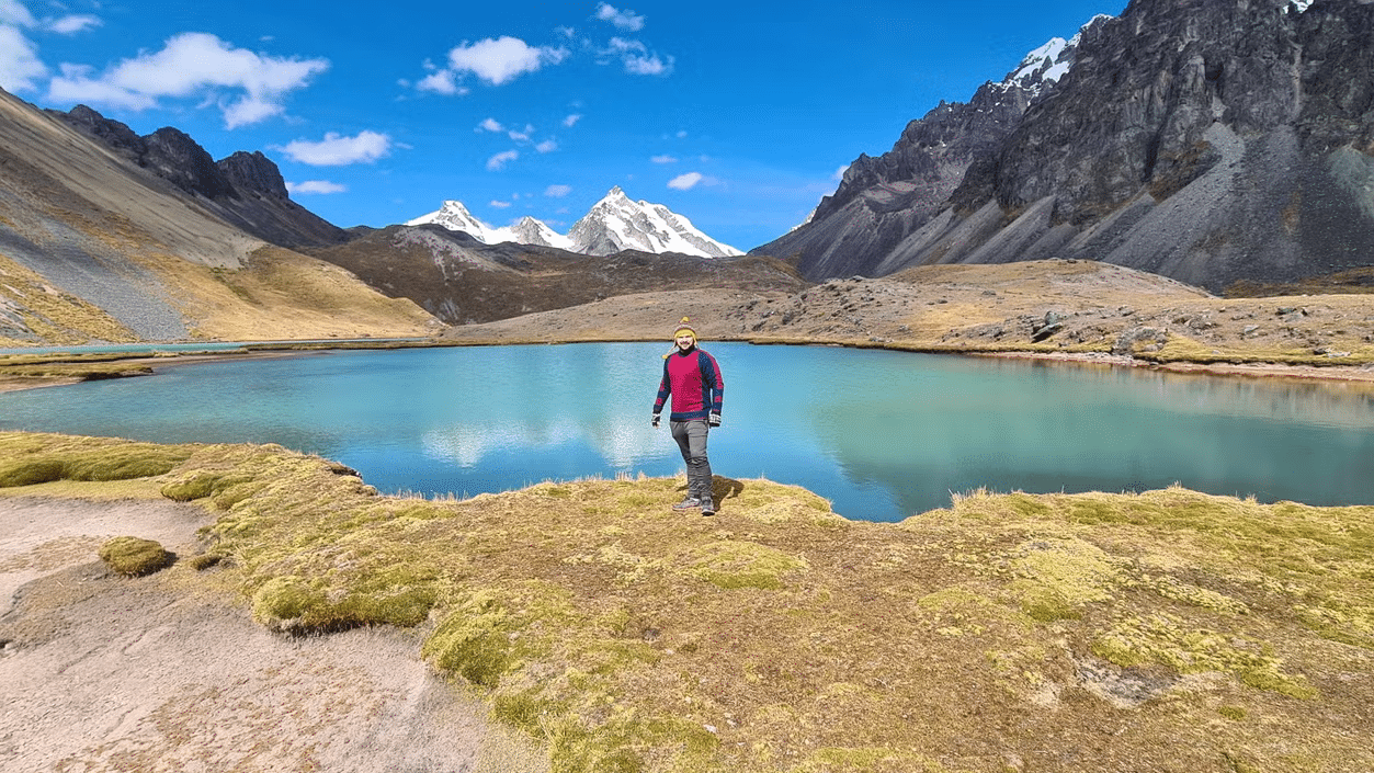 Homem de pé com uma lagoa cristalina ao fundo, rodeada de natureza e montanhas.