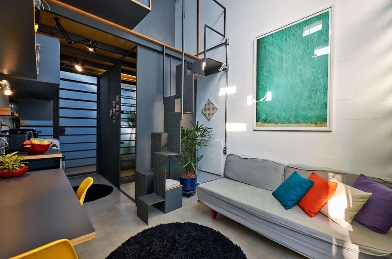 Aimagem retrata um espaço interno de um estúdio da Vila Pitanga, decorado de forma moderna e funcional. No canto esquerdo, uma cozinha compacta com armários e bancada em tons de cinza e amarelo, equipada com utensílios de cozinha e uma tigela de frutas. Ao centro, há uma escada de metal preta com degraus que parecem flutuar, levando a um segundo nível não visível na imagem. No canto direito, há um sofá de canto cinza claro com almofadas coloridas. Um grande quadro com uma fotografia de água em tons de azul turquesa adorna a parede branca acima do sofá. No chão, um tapete preto circular adiciona textura ao espaço. O ambiente transmite um ar contemporâneo e despojado.