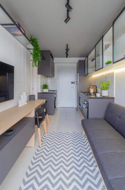 A imagem mostra o "O Melhor Stúdio da região!", um dos airbnb em São Paulo, moderno e estiloso. À direita, há um sofá cinza sobre um tapete com padrão em zigue-zague. Do lado esquerdo, uma cozinha planejada com armários superiores cinza escuro, bancada de trabalho clara e cadeiras de bar pretas e de madeira. Sobre a bancada, a cozinha está equipada com modernos eletrodomésticos e plantas decorativas. Há uma TV na extremidade da bancada. Luminárias de trilho no teto iluminam o espaço, e uma porta branca ao fundo indica a entrada do apartamento. O chão é de azulejo claro, contribuindo para a sensação de um ambiente limpo e aberto.