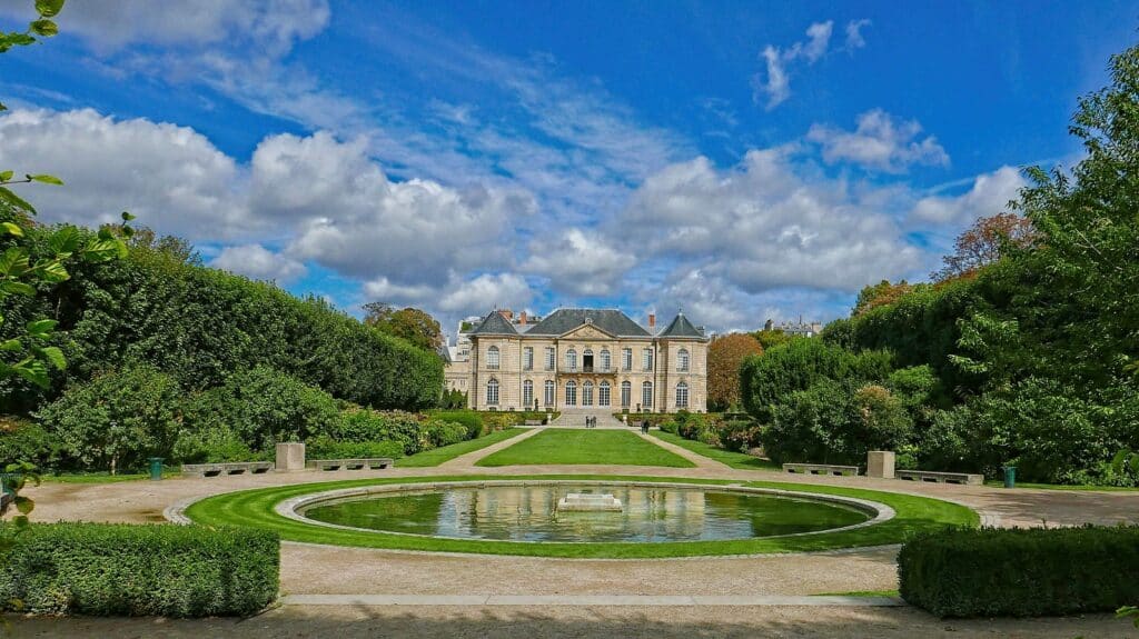 Uma fonte está no centro do jardim, que é cercado por árvores e gramados. Ao fundo fica o Hotel Biron, instalação onde funciona o Museu Rodin, uma das opções de o que fazer em Paris.