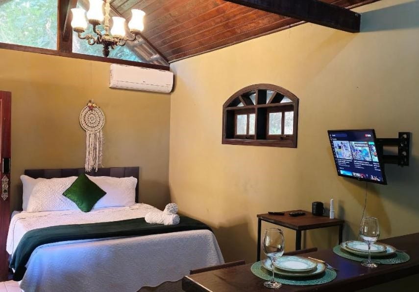 Quarto aconchegante da KORU Cabana. Há uma cama no lado esquerdo, TV na parede, ar condicionado, uma pequena área de jantar para dois e um apanhador de sonhos acima da cama. Esse é um dos bons chalés em Petrópolis.